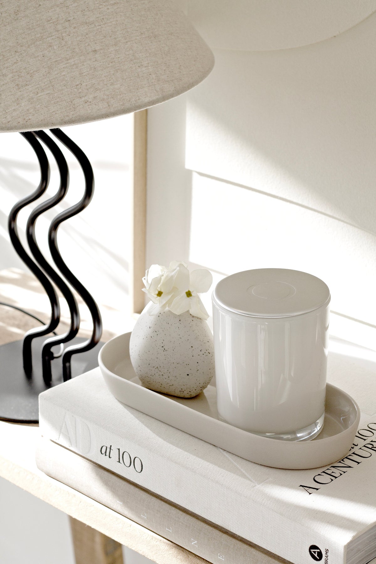 Sunlit side-table vignette with a wavy black lamp, speckled bud vase of white flowers, and a lidded white candle on a ceramic tray atop design books