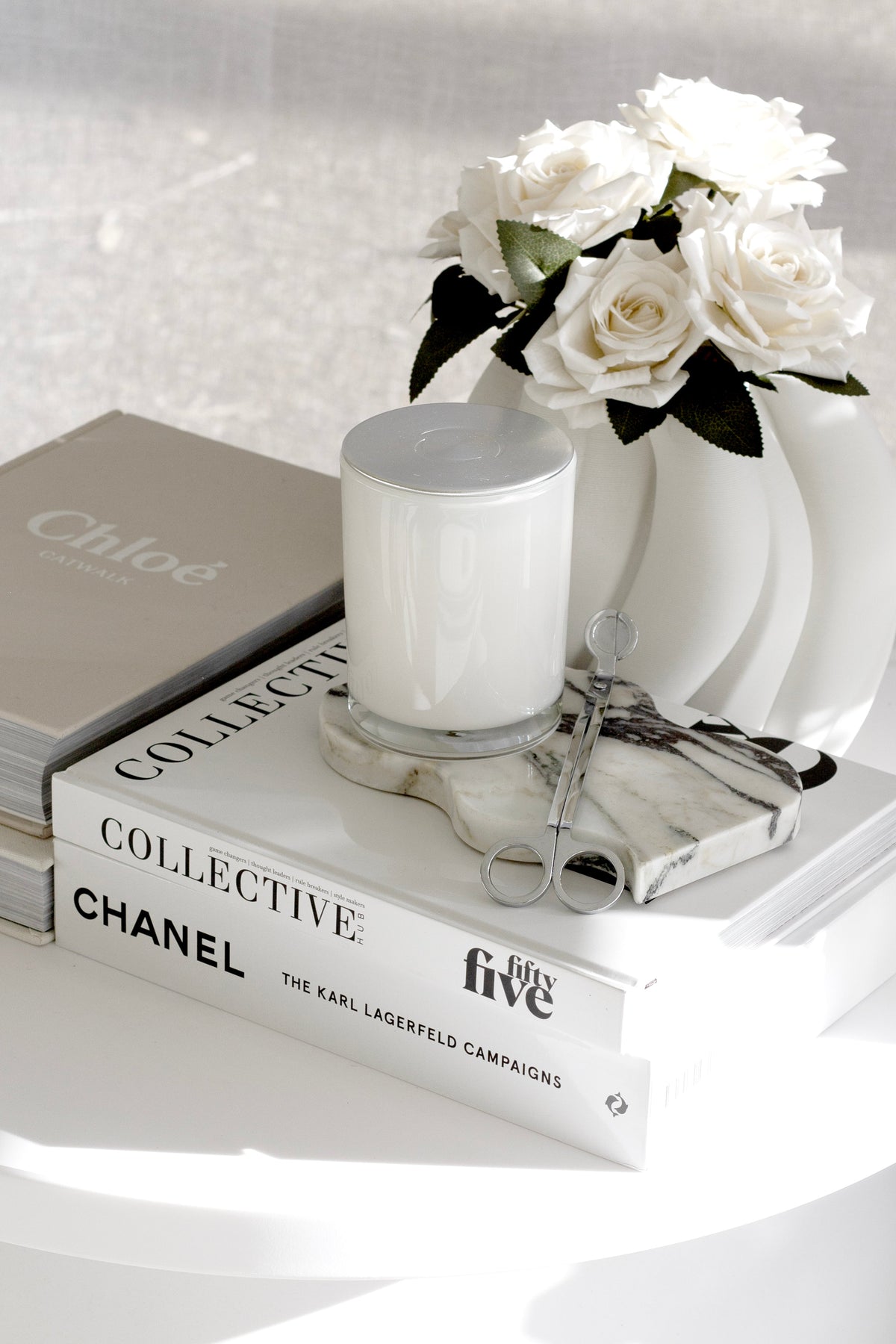Candle and silver candle tools on a wavy marble tray beside white roses on books