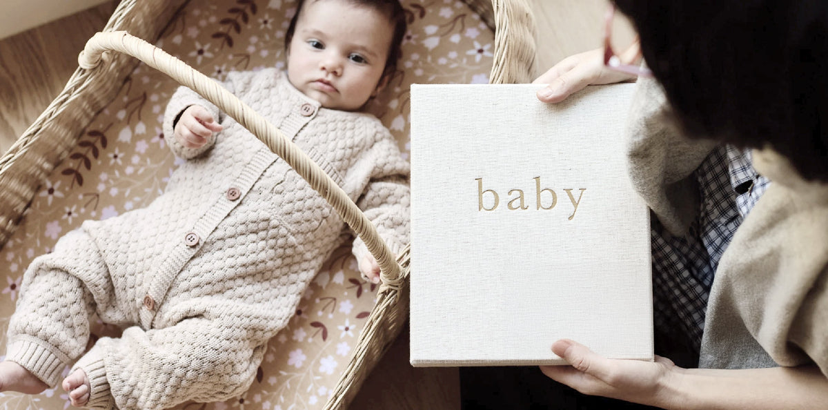 Baby lying in a woven basket beside a hand holding a linen Write To Me baby journal
