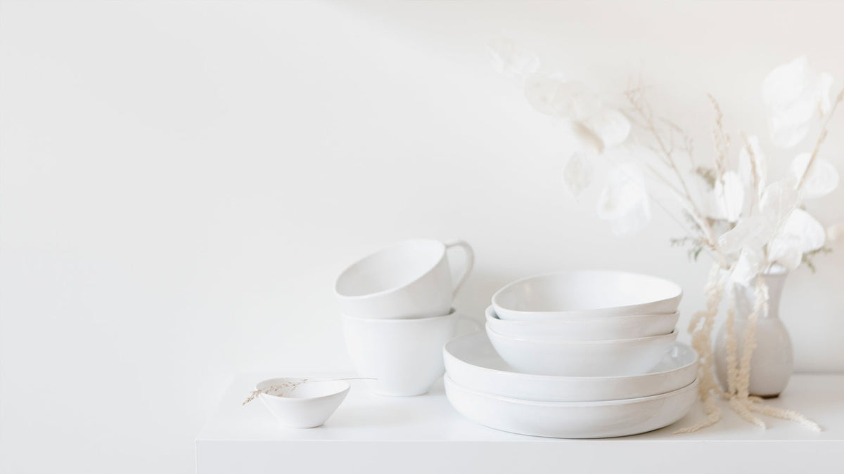 Stacked white ceramic bowls and plates with two cups and a small pinch bowl beside a vase of dried white foliage on a white shelf