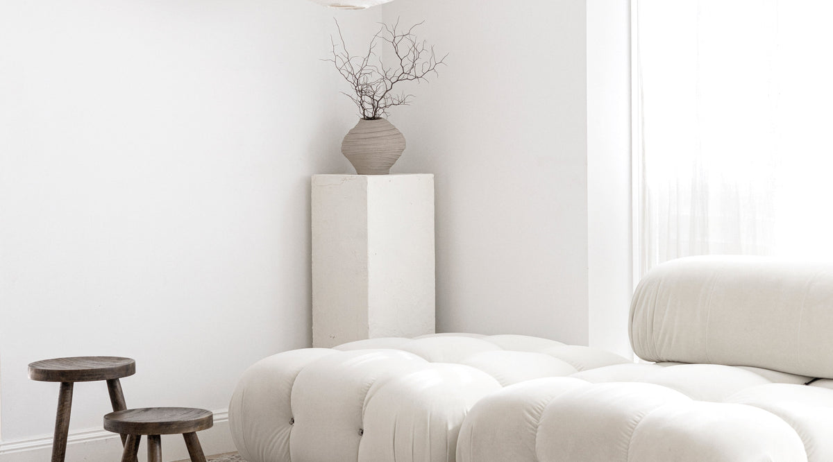Minimal living room with a modular cream sofa, a timber stool and a vase of branches on a plinth