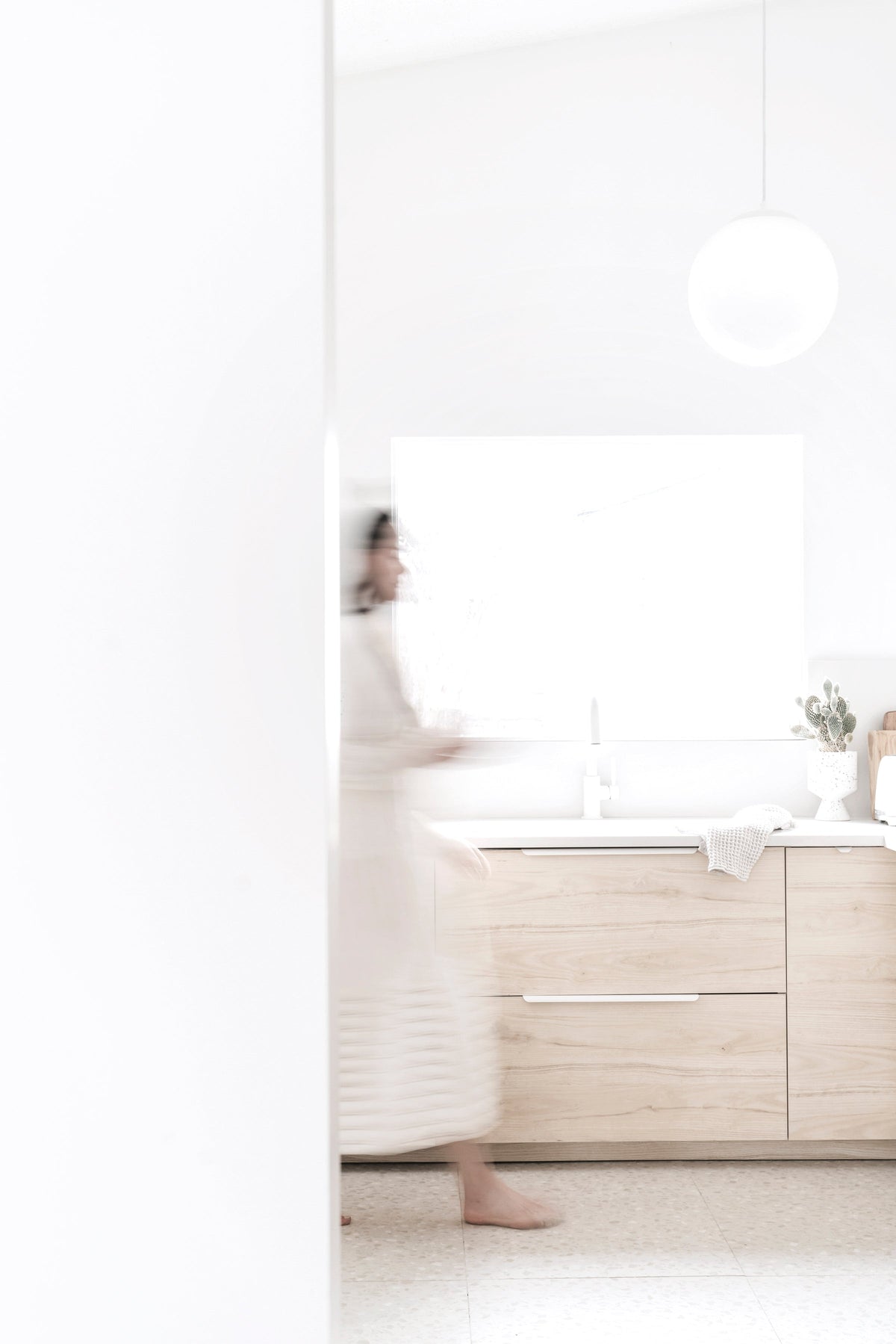 Person in a white robe walking through a bright kitchen with pale timber cabinets