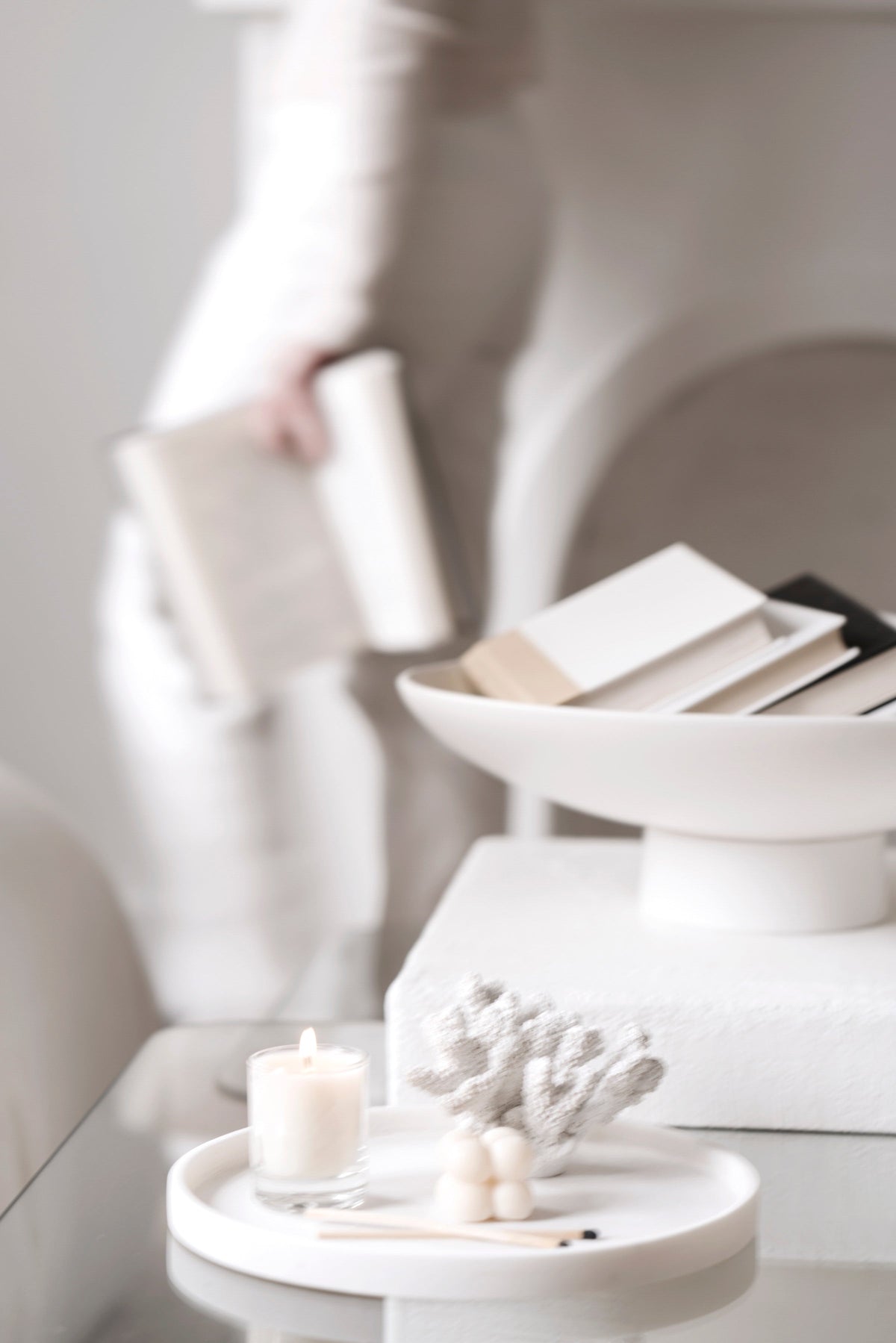 Person in cream reading beside a pedestal bowl of books, a candle and coral on a white side table
