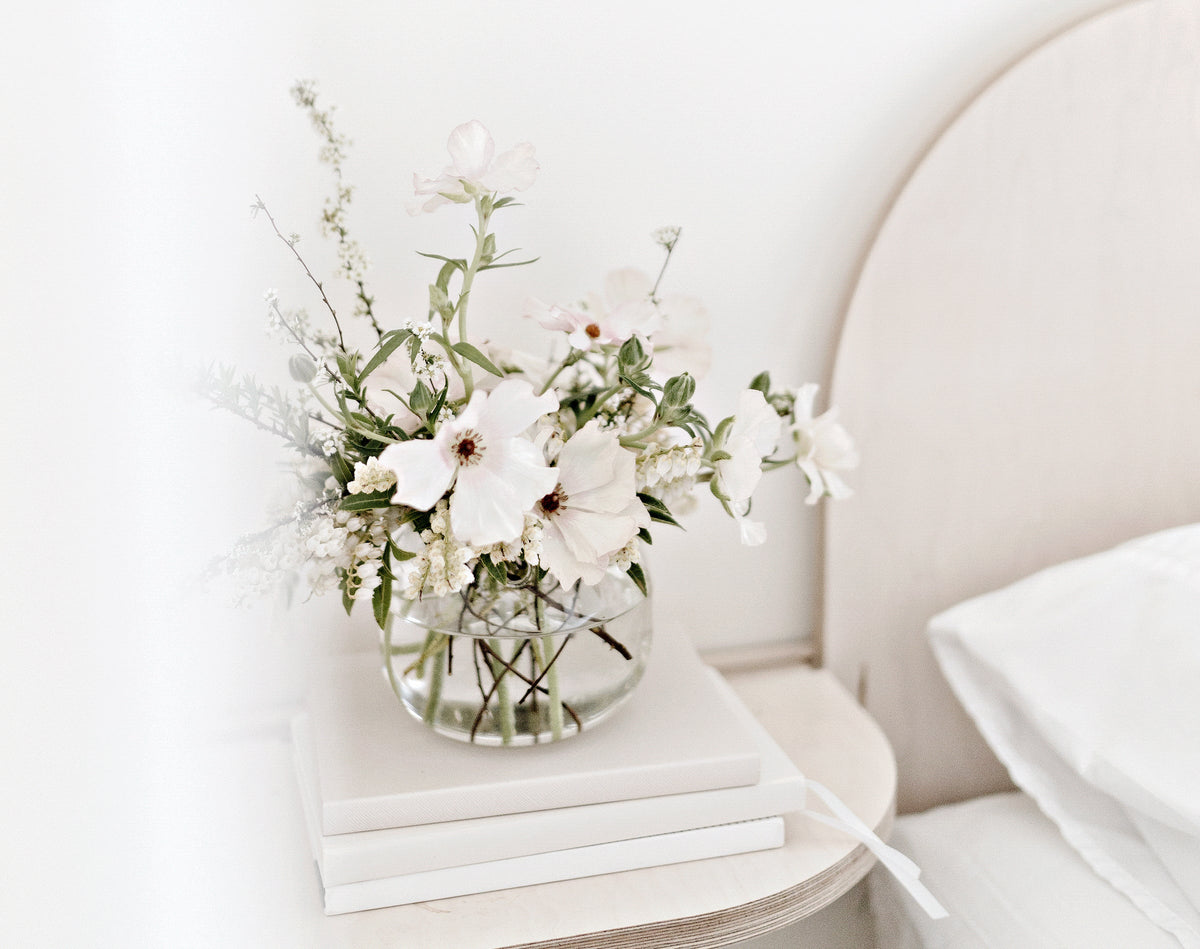 Glass vase of white flowers on a stack of books beside an upholstered bed