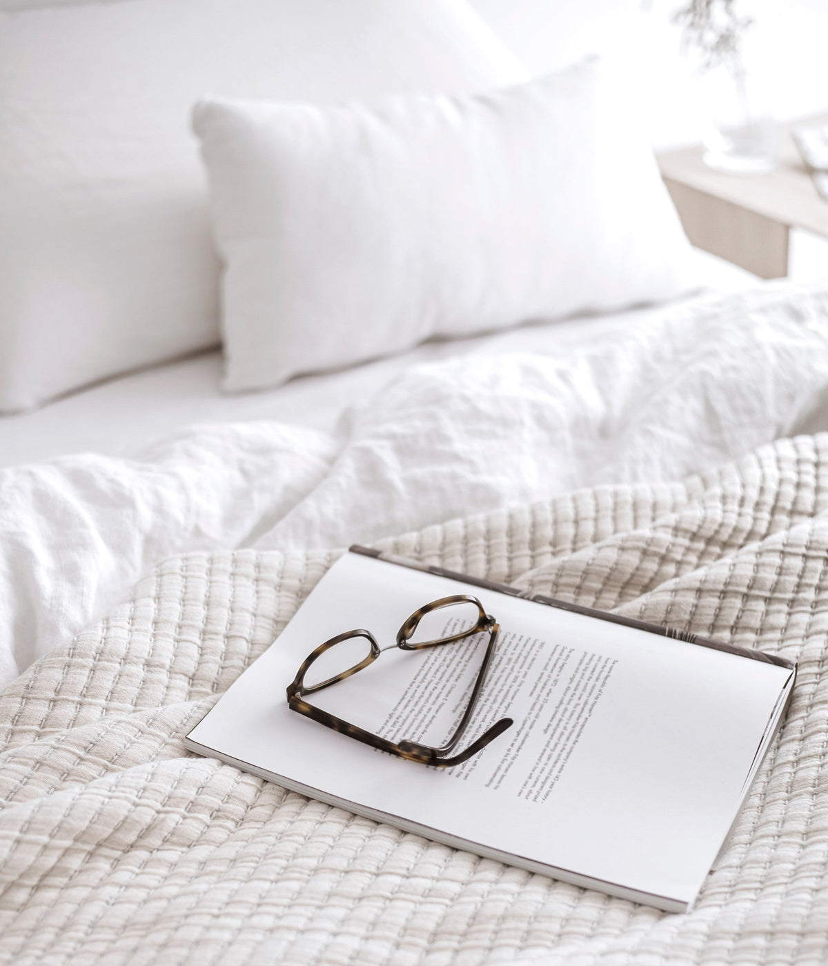 Neutral bed layered with white and cream linens, an open book and reading glasses