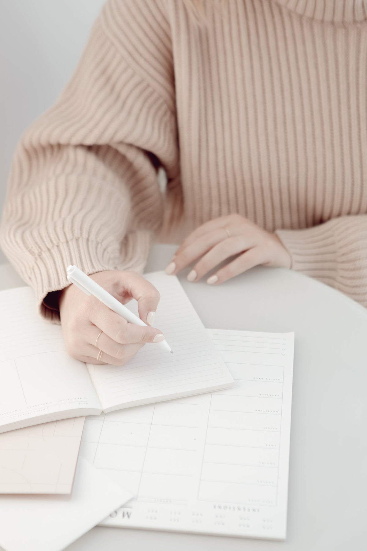 Hands in a blush knit writing in a notebook on a white desk