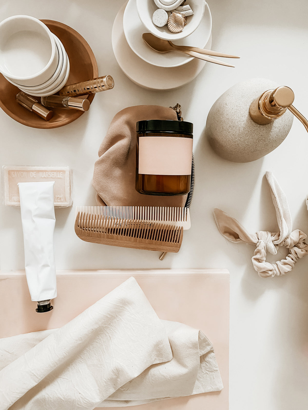 Flat lay of hand cream, a comb, a candle, a scrunchie and linen in soft blush tones