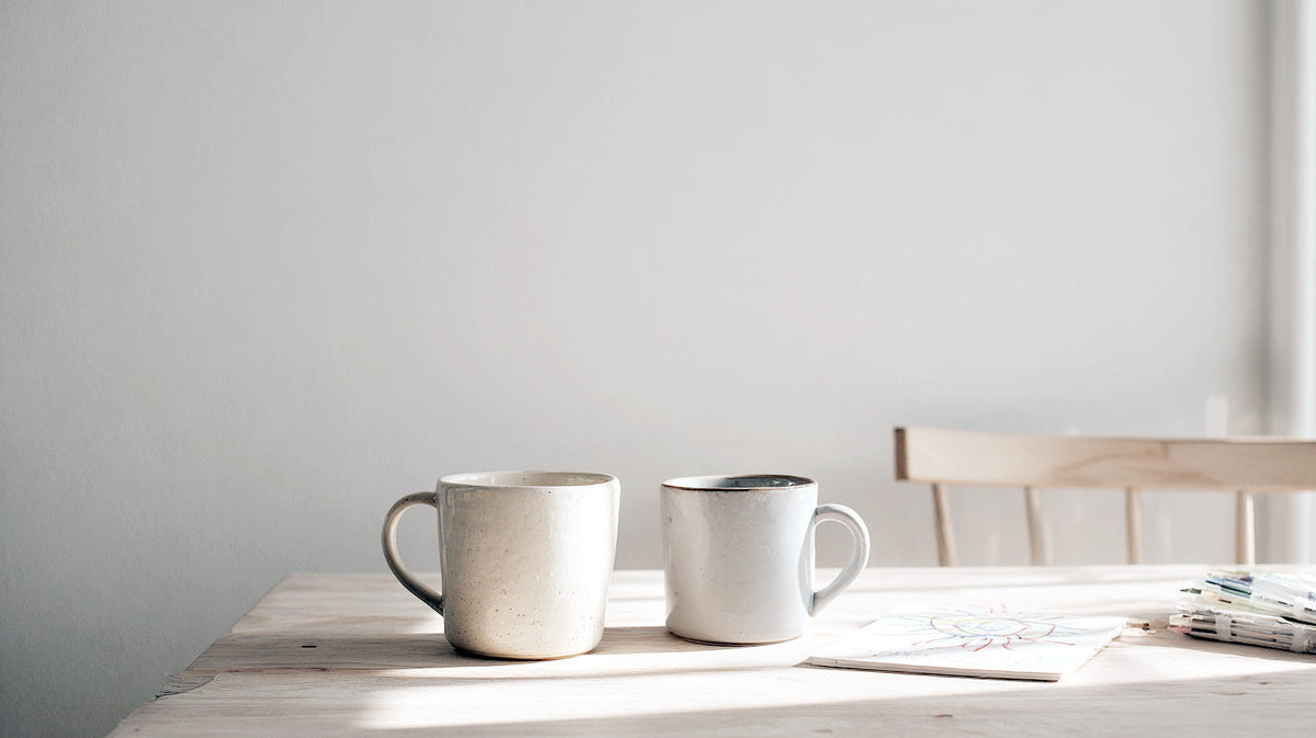 Two white mugs on a pale timber table in soft daylight