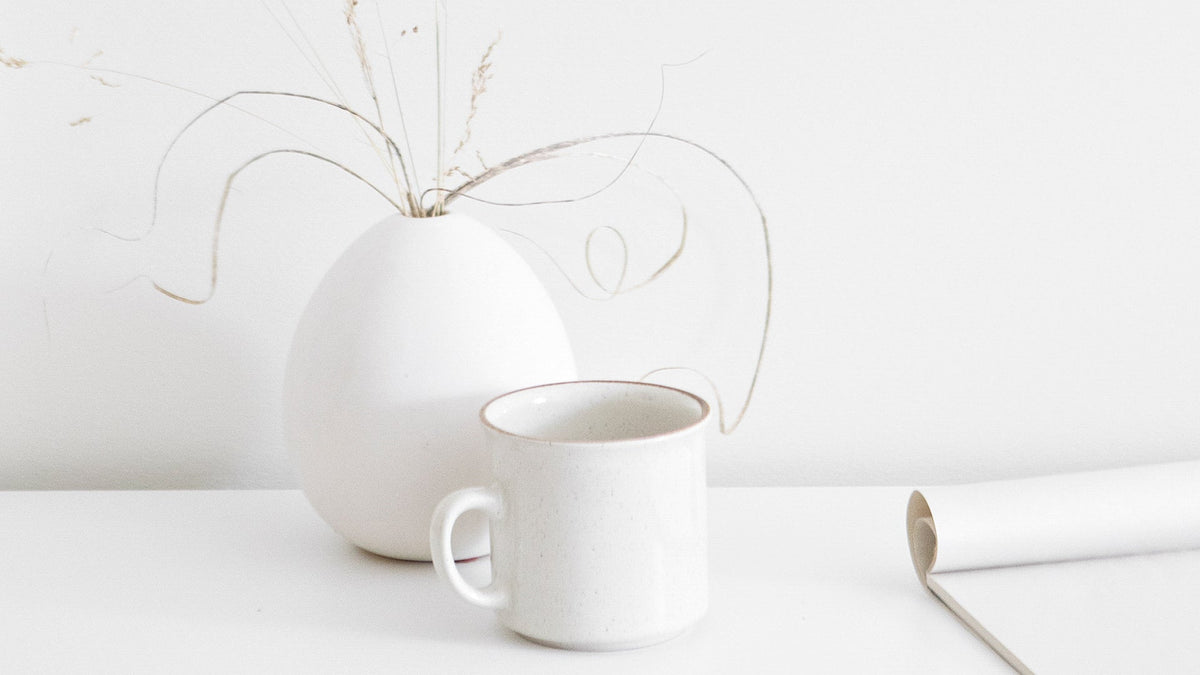 Minimalist white ceramic mug and vase with dried grasses on a white surface, styled with calming simplicity to evoke softness, stillness, and nervous system regulation.