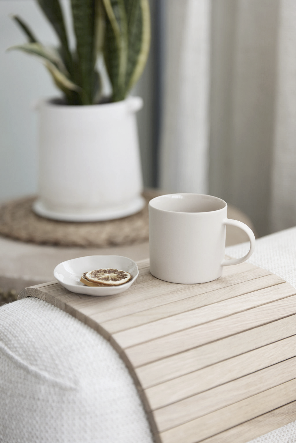Oak slatted armchair tray on a sofa arm with a ceramic mug and a small plate of dried lemon, a potted snake plant behind