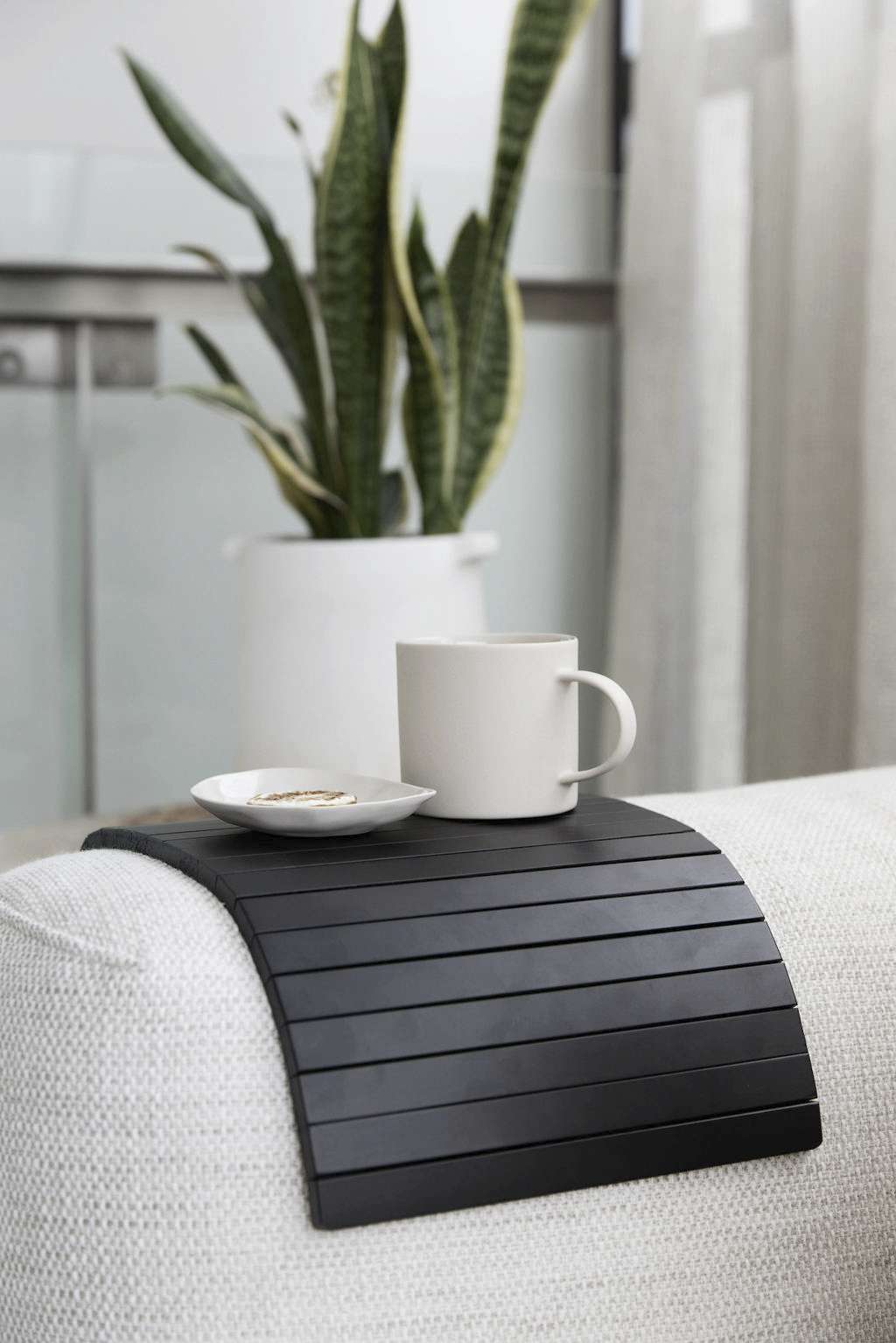 Black slatted armchair tray on a white sofa arm with a mug and plate, a potted snake plant behind