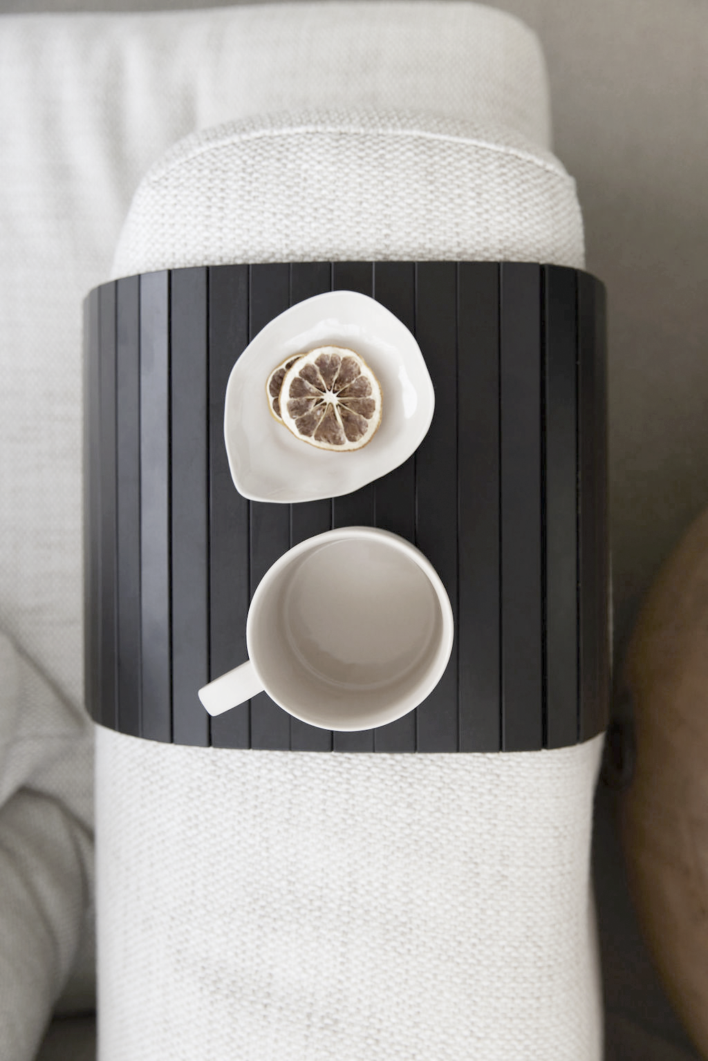 Black slatted armchair tray over a sofa arm holding a mug and a small plate with a slice of dried lemon