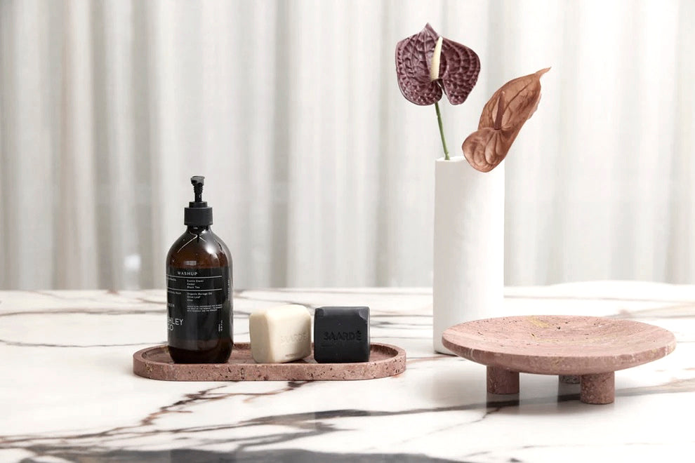 Burnt Travertine Nome stand with a pump bottle and small candle on a marble table beside anthurium stems