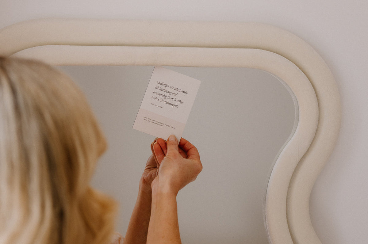 Woman holding an Art of Living card up to a curved cream mirror