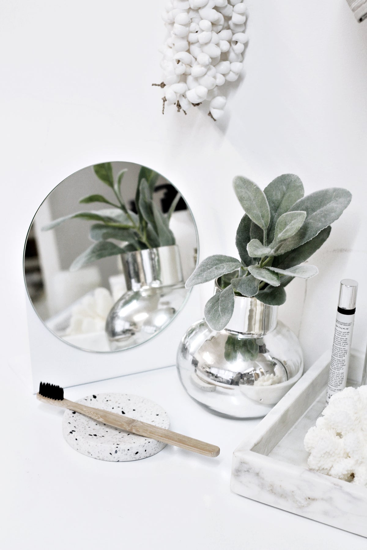 Grey marble tray on a white table beside a round mirror, silver bud vases and greenery