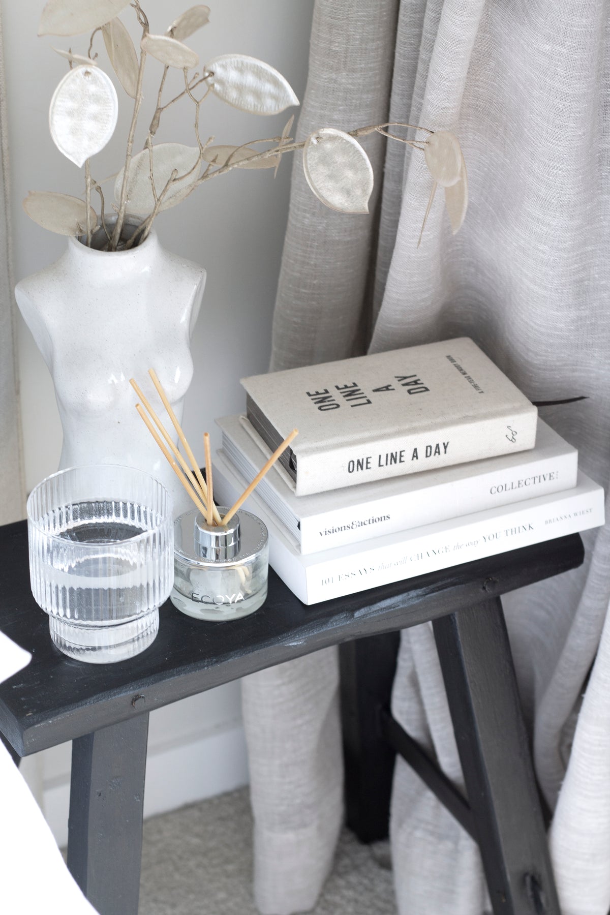 Female Silhouette Vase with dried stems on a black stool beside books and a glass