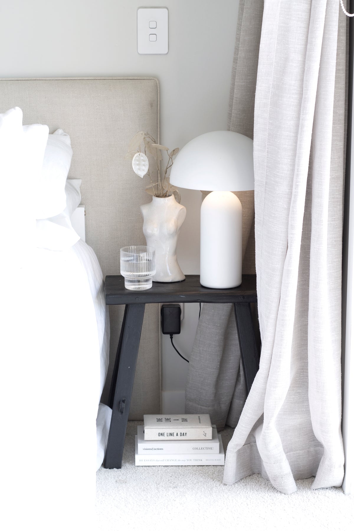 Black bench used as a bedside table with a white dome lamp, torso vase and books on the shelf below