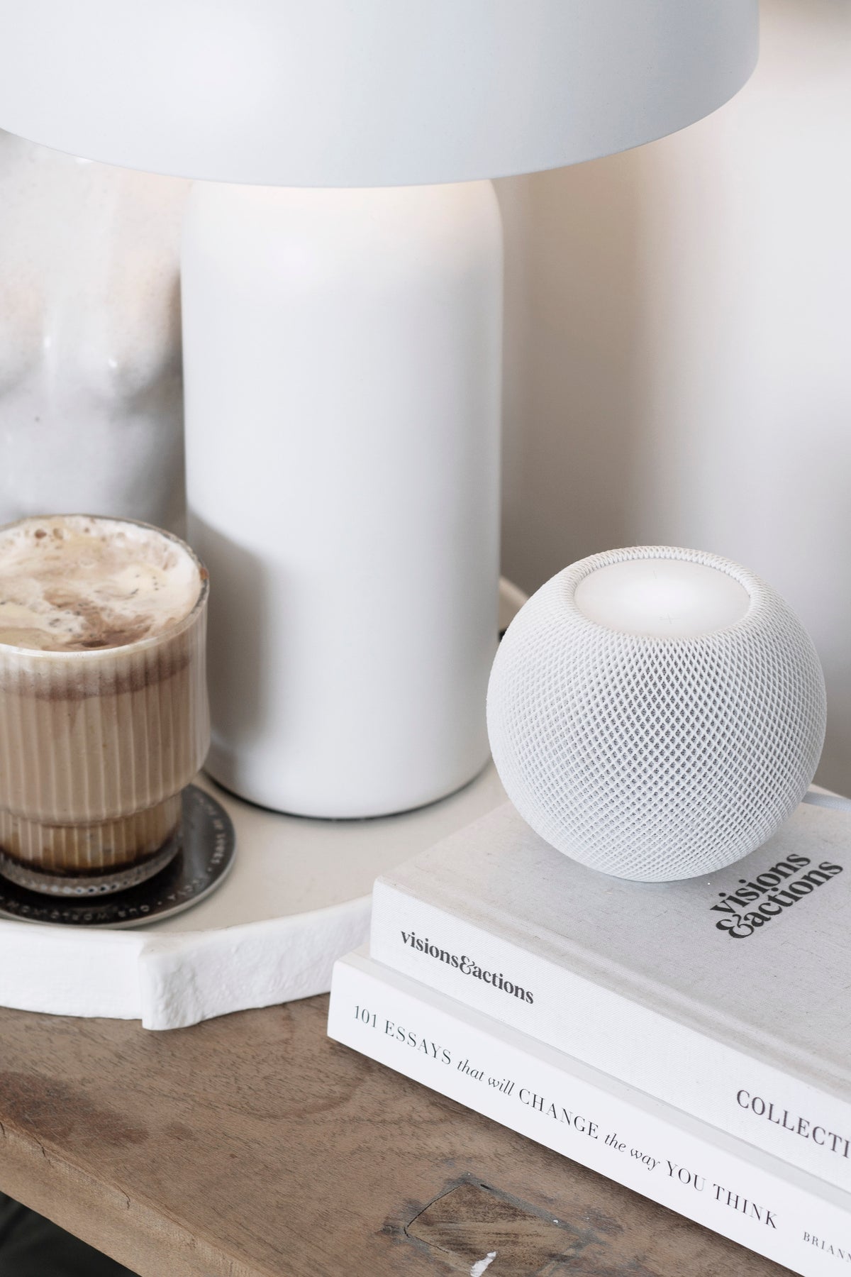 Bedside timber surface with a table lamp, a fluted candle, a speaker and a stack of books