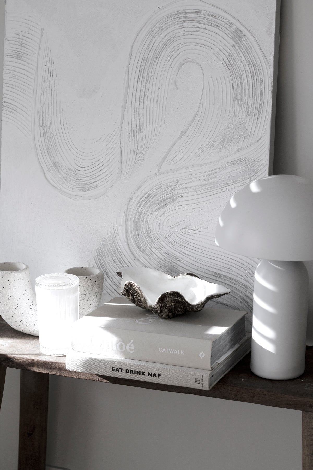 Clam shell dish on books beneath an abstract line artwork, with a ribbed vase and a dome lamp