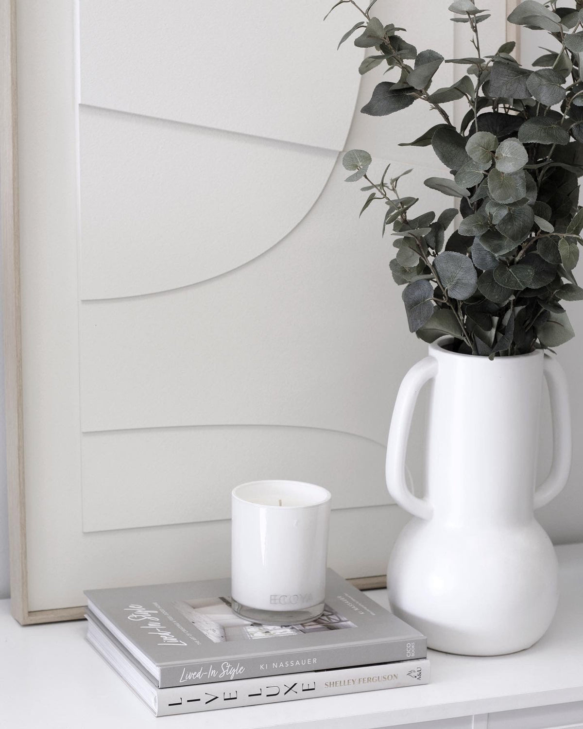 Faux eucalyptus stems in a white jug vase with books and a candle on a console