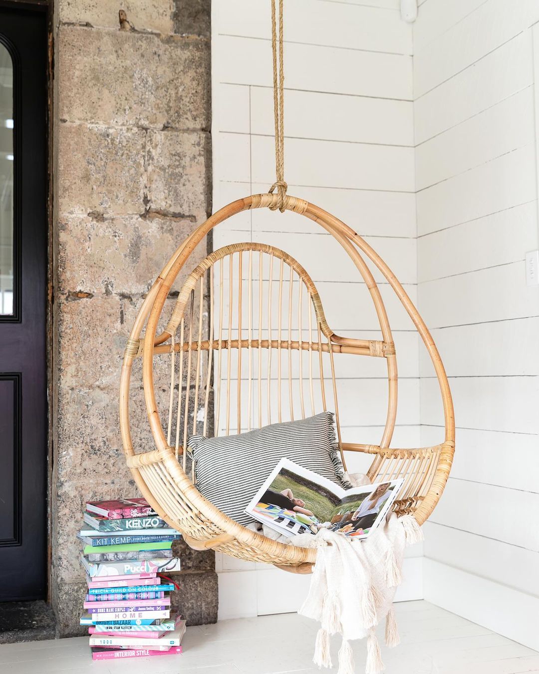 Rattan hanging chair suspended in a corner with a cushion, magazines and books below
