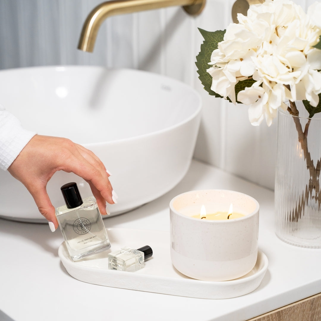 Hand setting a perfume oil bottle on a tray beside a lit candle and white hydrangeas