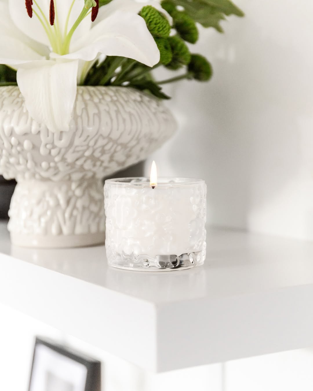 A lit FLWR candle in embossed glass on a white ledge, white lilies in a textured vase behind