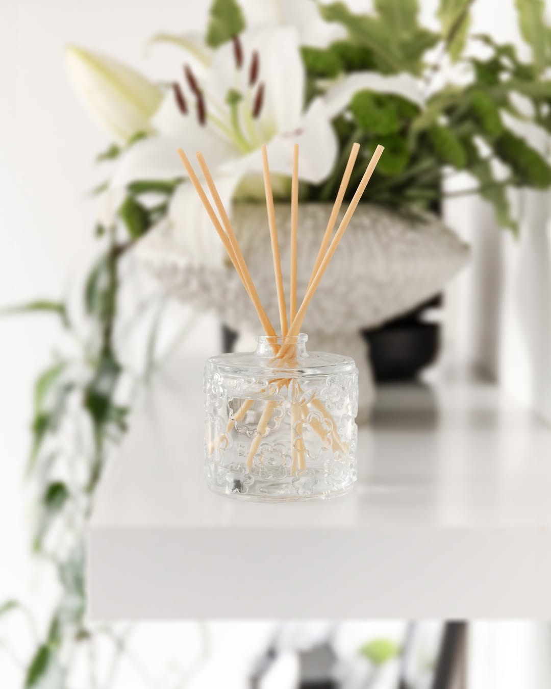 A FLWR reed diffuser in embossed glass among white lilies on a bright white ledge