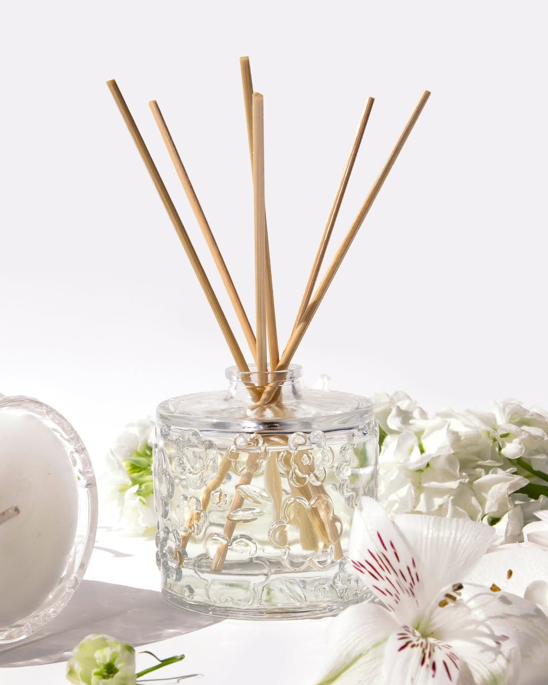 A FLWR reed diffuser with pale reeds, white blooms around it and a candle just behind