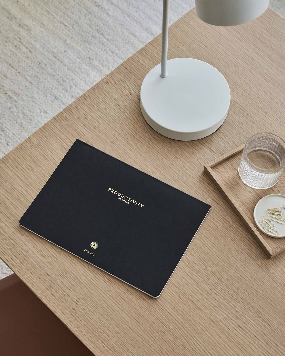 Productivity desk pad on a timber desk beside a lamp, a glass and a small dish