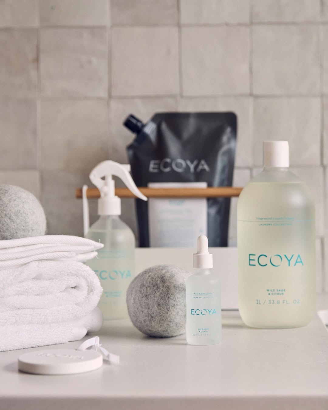 Stack of white towels with Ecoya dryer balls, linen spray, refill pouch and laundry liquid against tiles