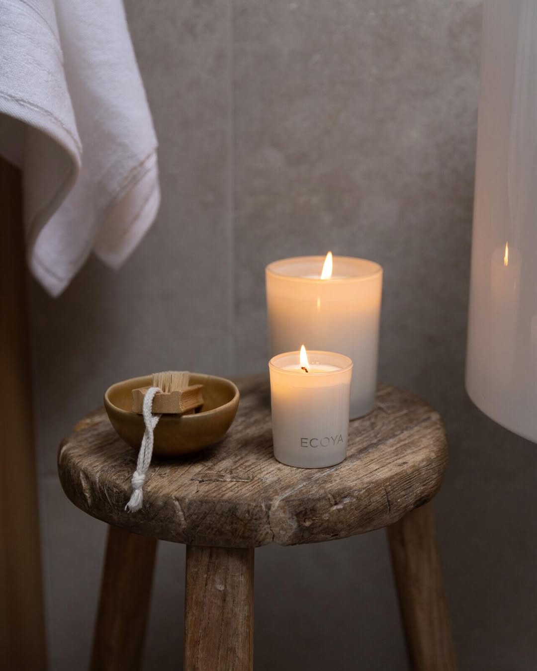 Two lit Ecoya Madison candles on a rustic wooden stool in a bathroom, beside a small wooden bowl and a hanging towel