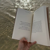 A person holding an open book, "This Was Meant To Find You (When You Needed It Most)" by Thought Catalog, in front of a body of water.