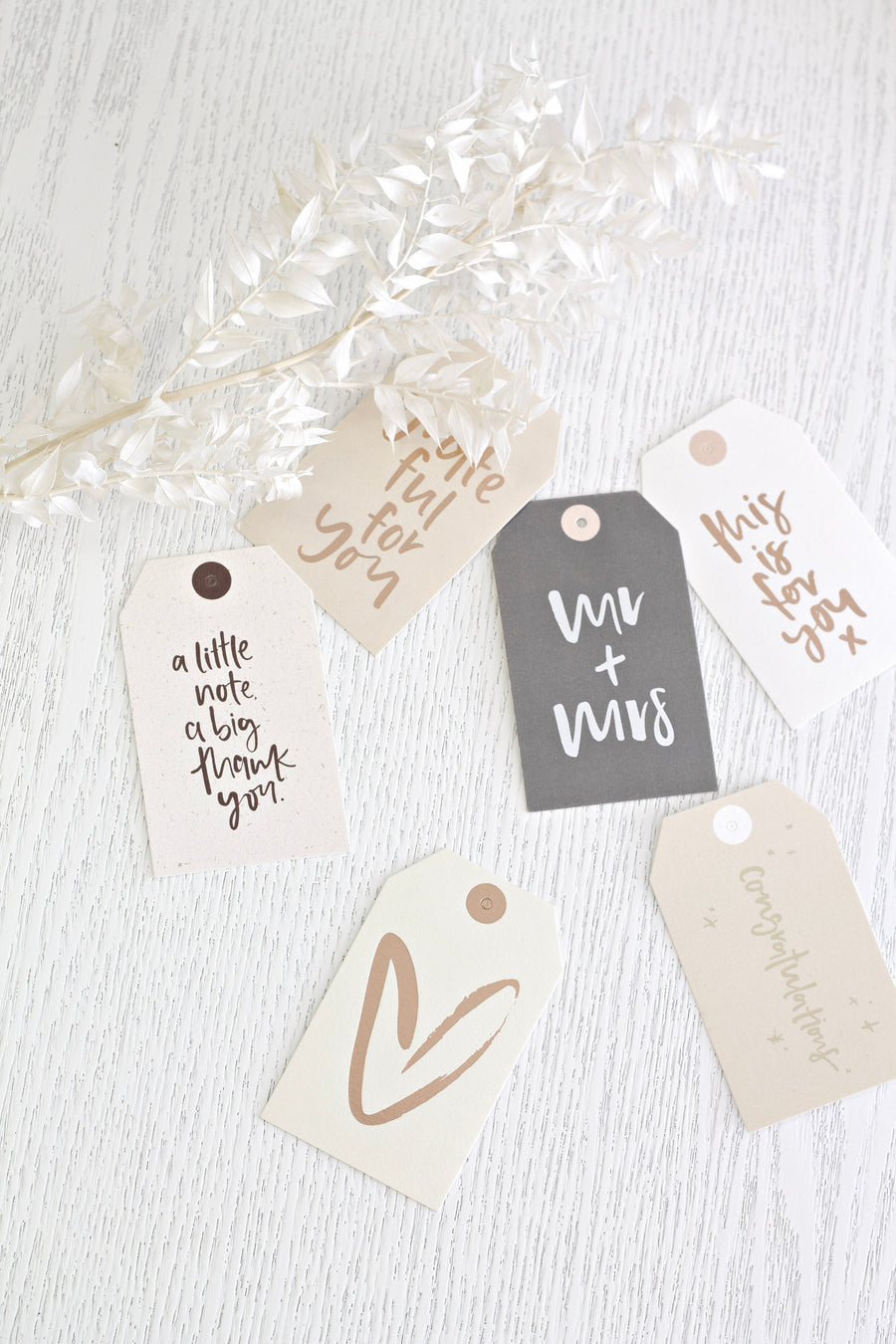 Several gift tags in cream and charcoal arranged on white linen with sprigs of dried white blossom