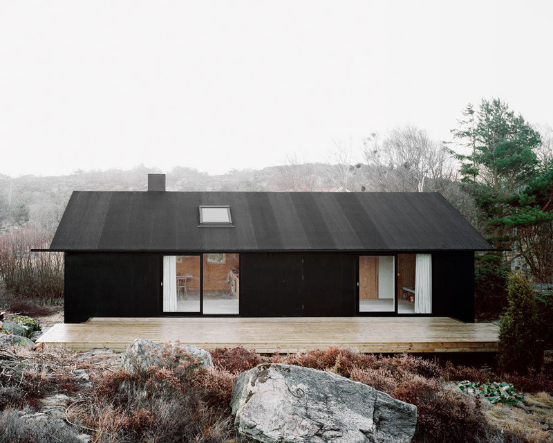 A black cabin, inspired by the lifestyle books "Rock the Shack" from Books, sits on top of rocks in the woods.