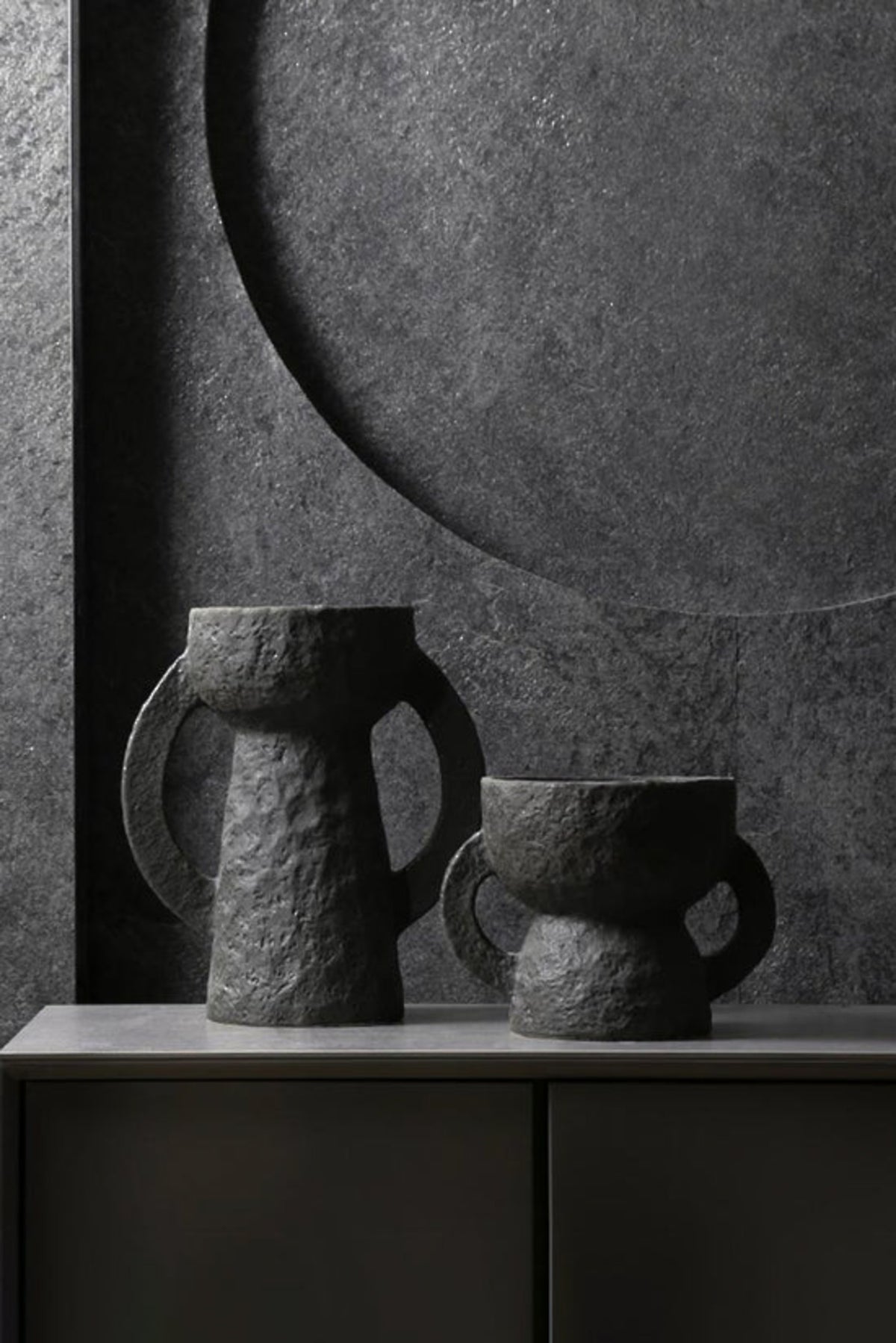 Large and small charcoal amphora vases on a dark sideboard against a textured grey wall