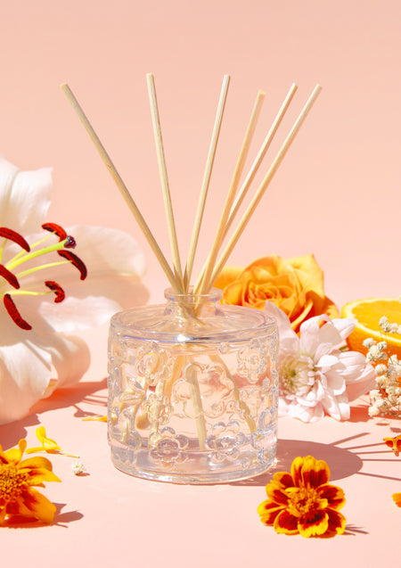 The Fleur D'Oranger diffuser on a peach backdrop with lilies, marigolds and an orange rose