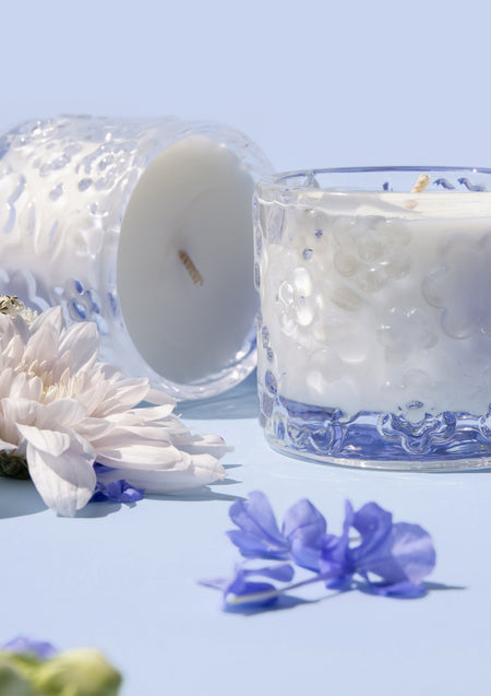 Two Forget Me Not candles, one on its side, among white dahlias and blue petals on pale blue
