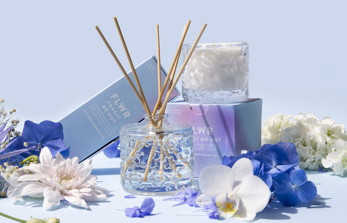 The Forget Me Not diffuser, candle and carton among white orchids and blue blooms