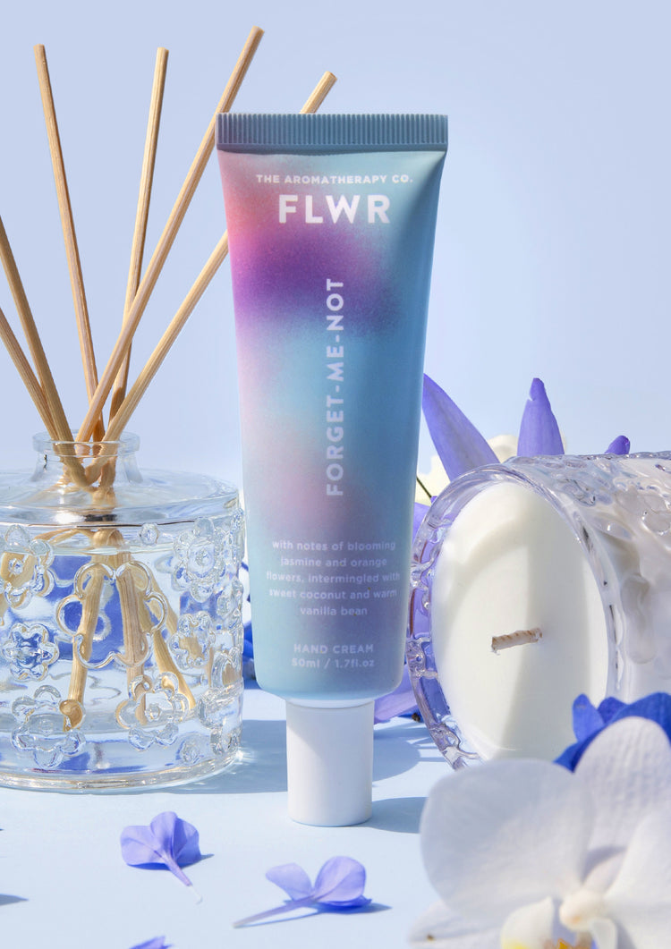 Forget-Me-Not hand cream tube with a glass reed diffuser, candle and white orchid on pale blue