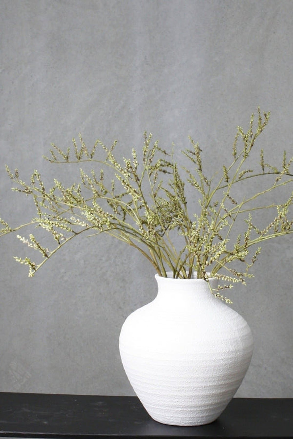 Tall branching stems with tiny pale buds in a ribbed white urn vase on a dark bench against a grey wall