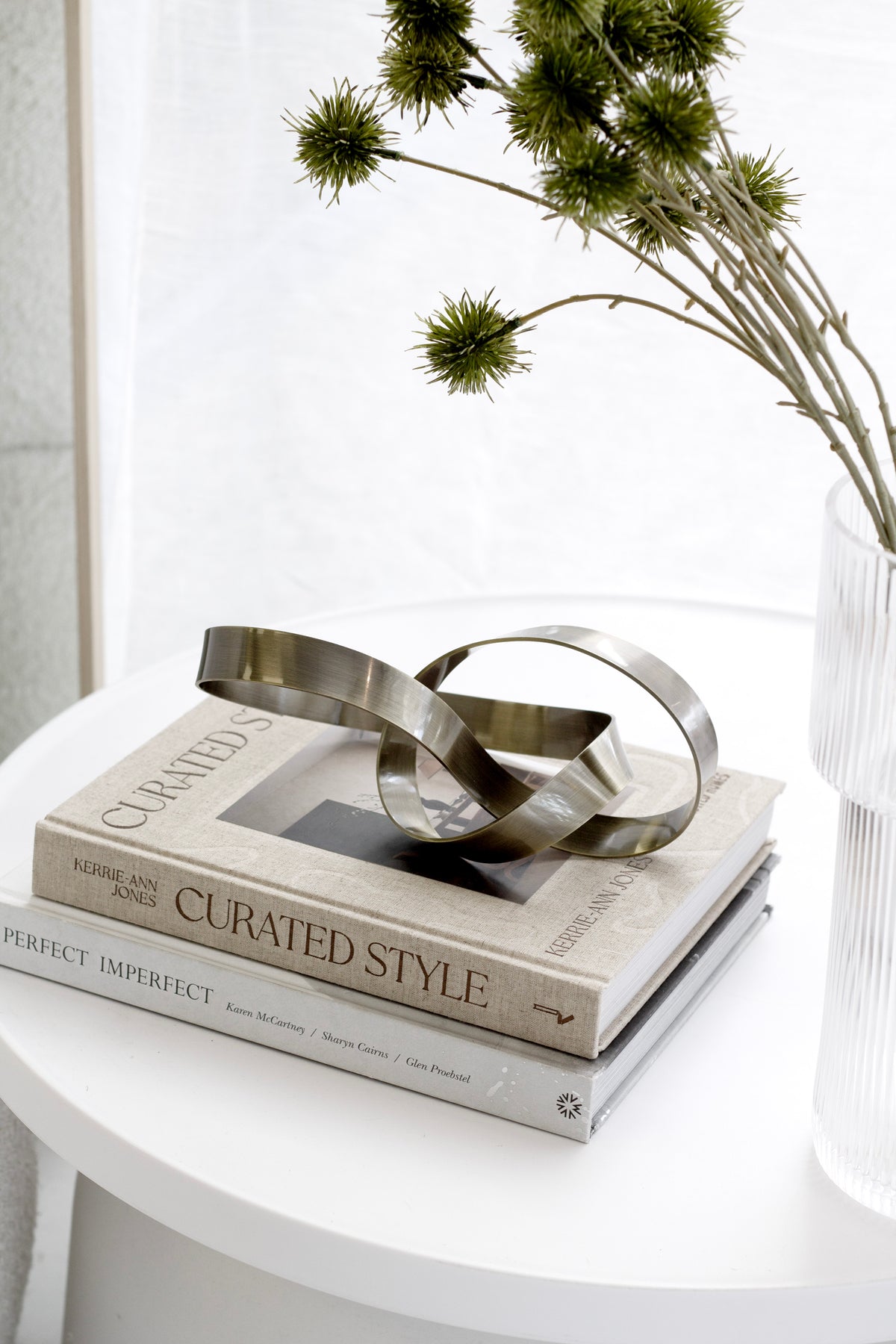 Bronze Infinity Loop sculpture on stacked books on a round white side table, with eryngium in a ribbed vase