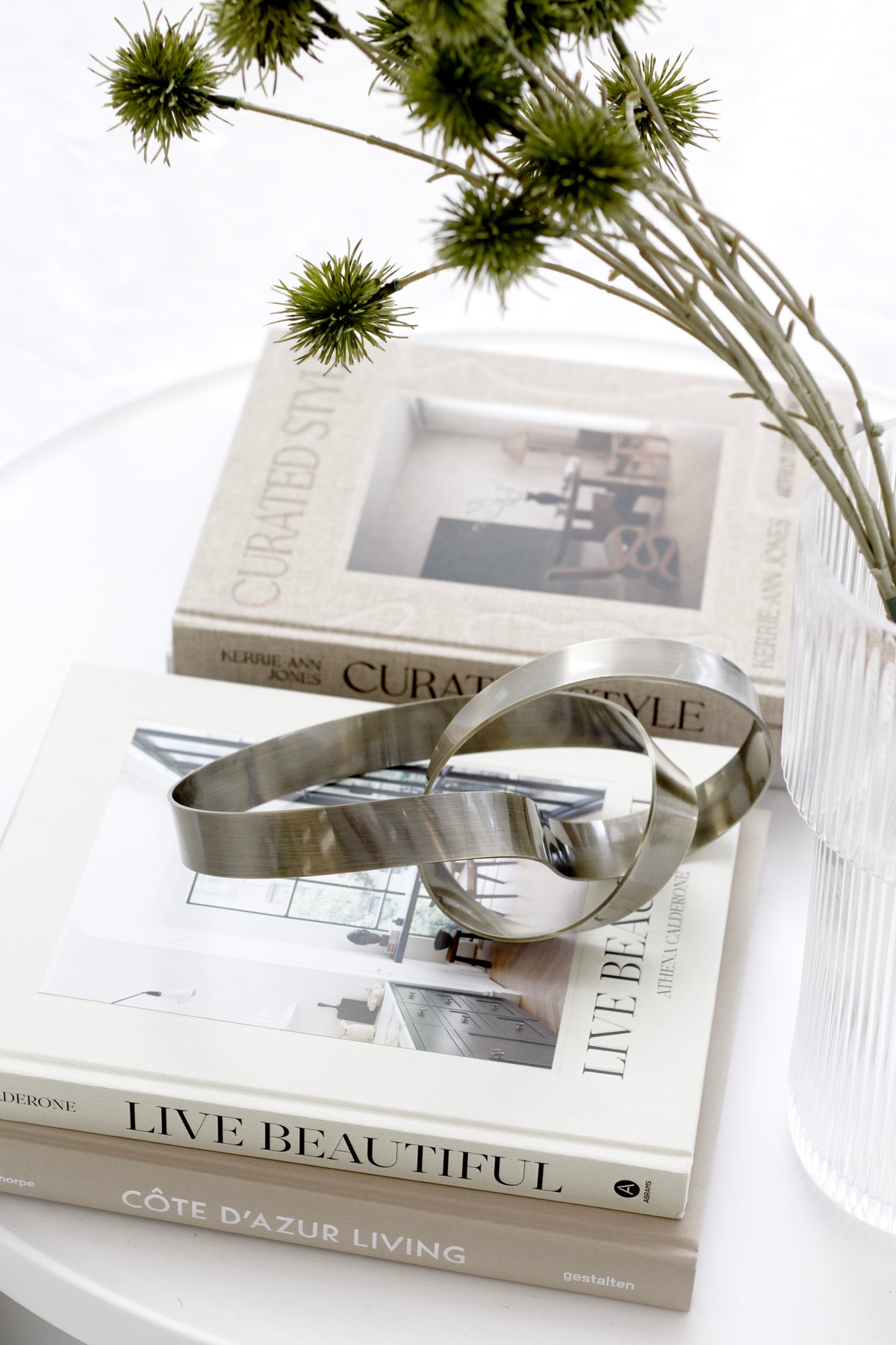 Bronze Infinity Loop sculpture on interiors books beside dried eryngium stems in a ribbed glass vase