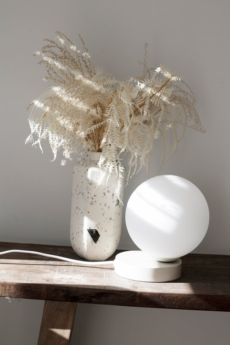 A vase of dried flowers sits on a wooden table next to a soft glow from a Yuri Table Lamp by Ned Collections.
