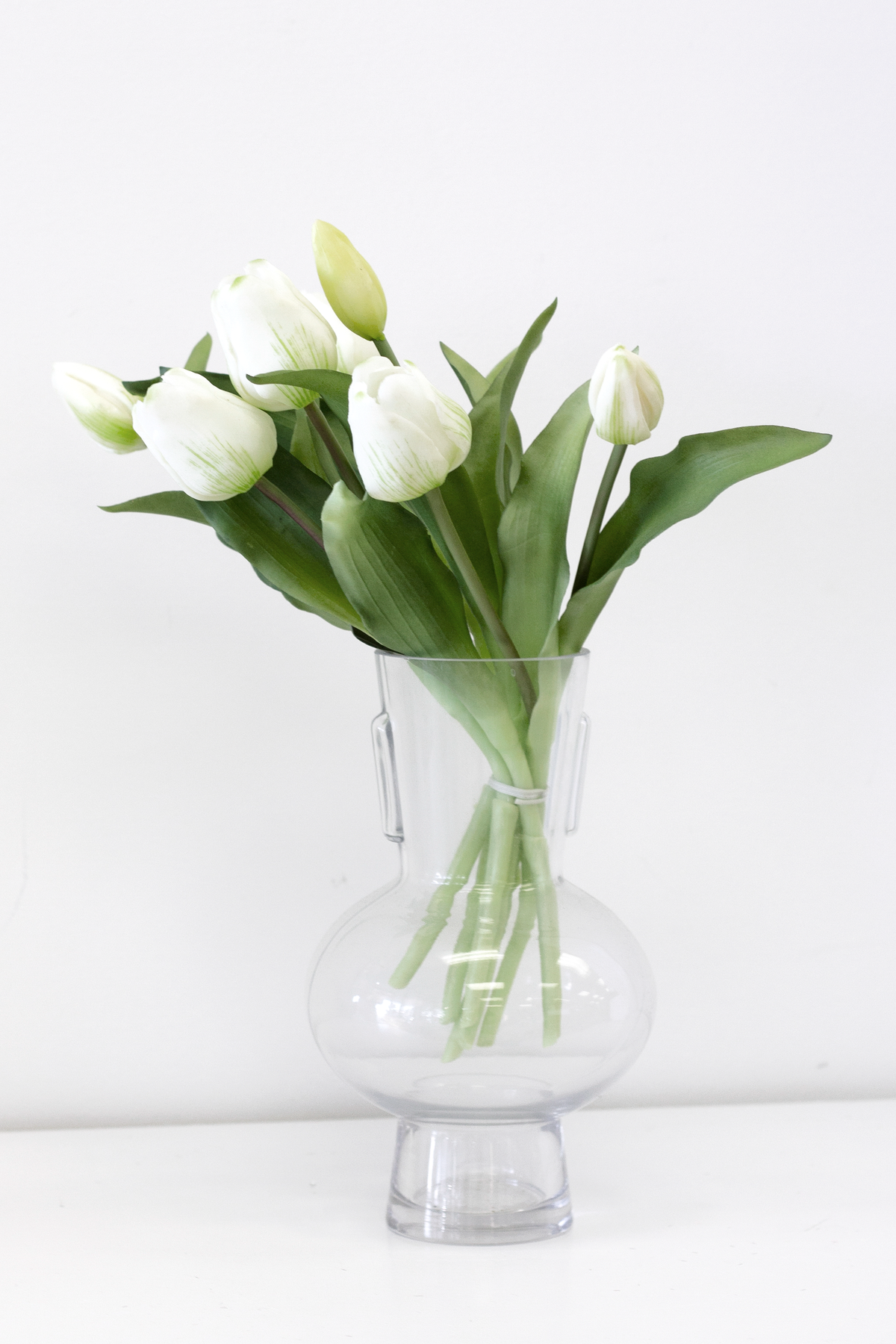 Tall white faux tulips in a round glass vase, front view on white