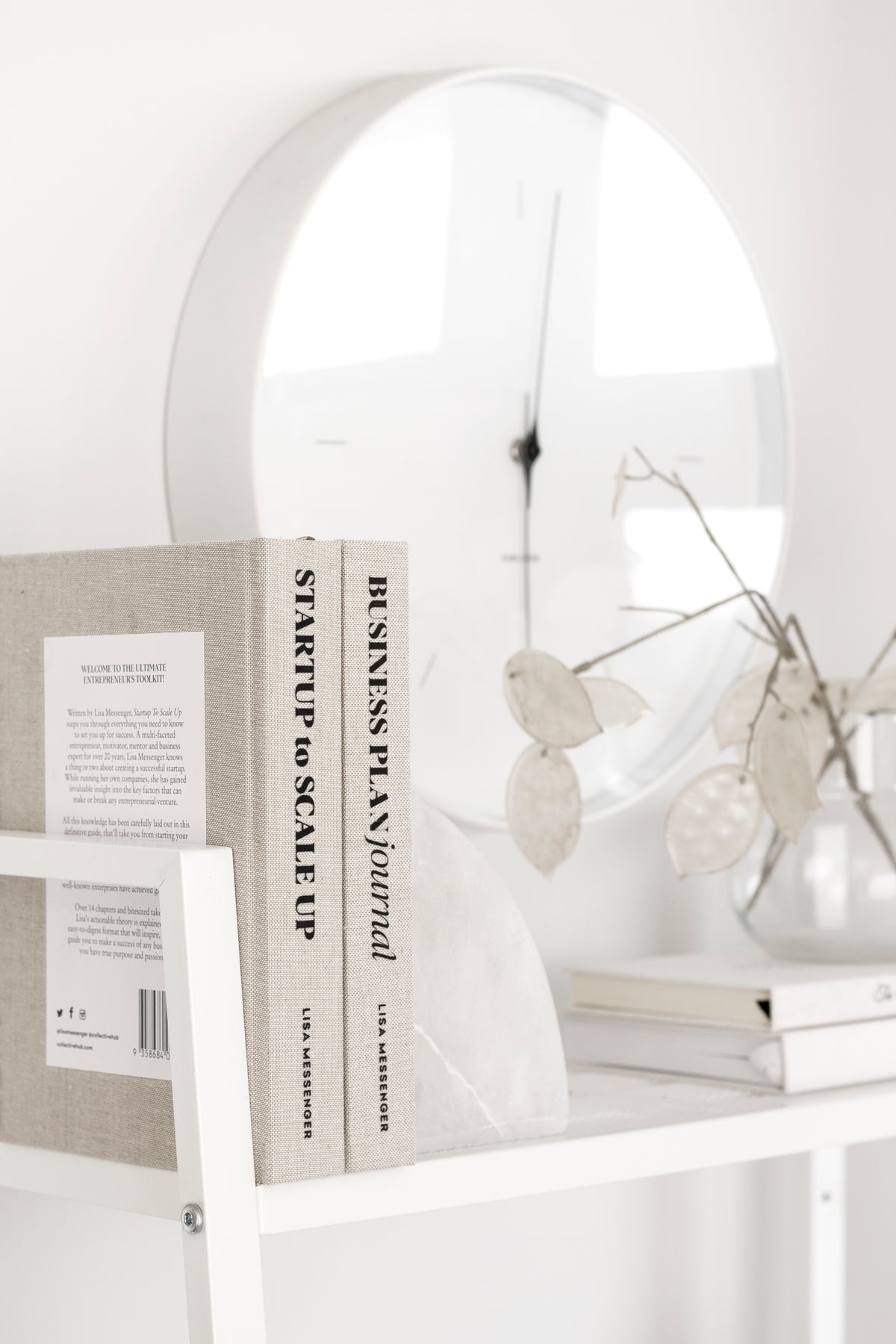 Close-up of two neutral-toned linen journals held by a curved white marble bookend on a modern white shelf, with a clear vase of dried lunaria and a sleek wall clock in the background.