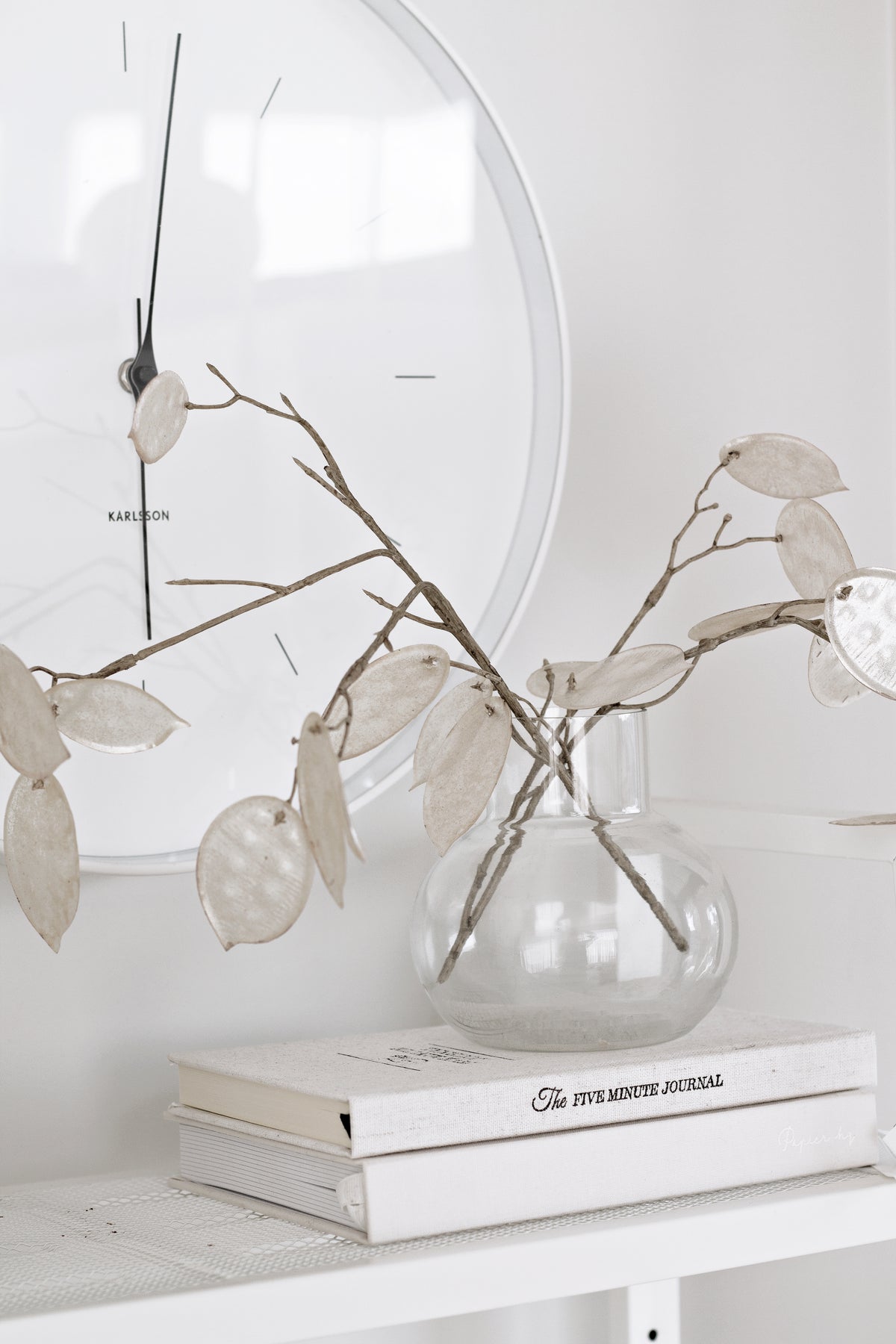 Five Minute Journal on a white shelf below a glass vase of dried honesty and a round mirror