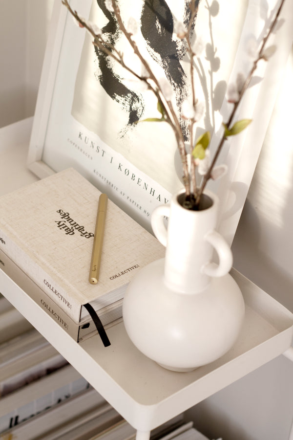 A white shelf filled with Papier HQ Brass Reusable Pens and limited edition books and stationery, adorned with a vase.