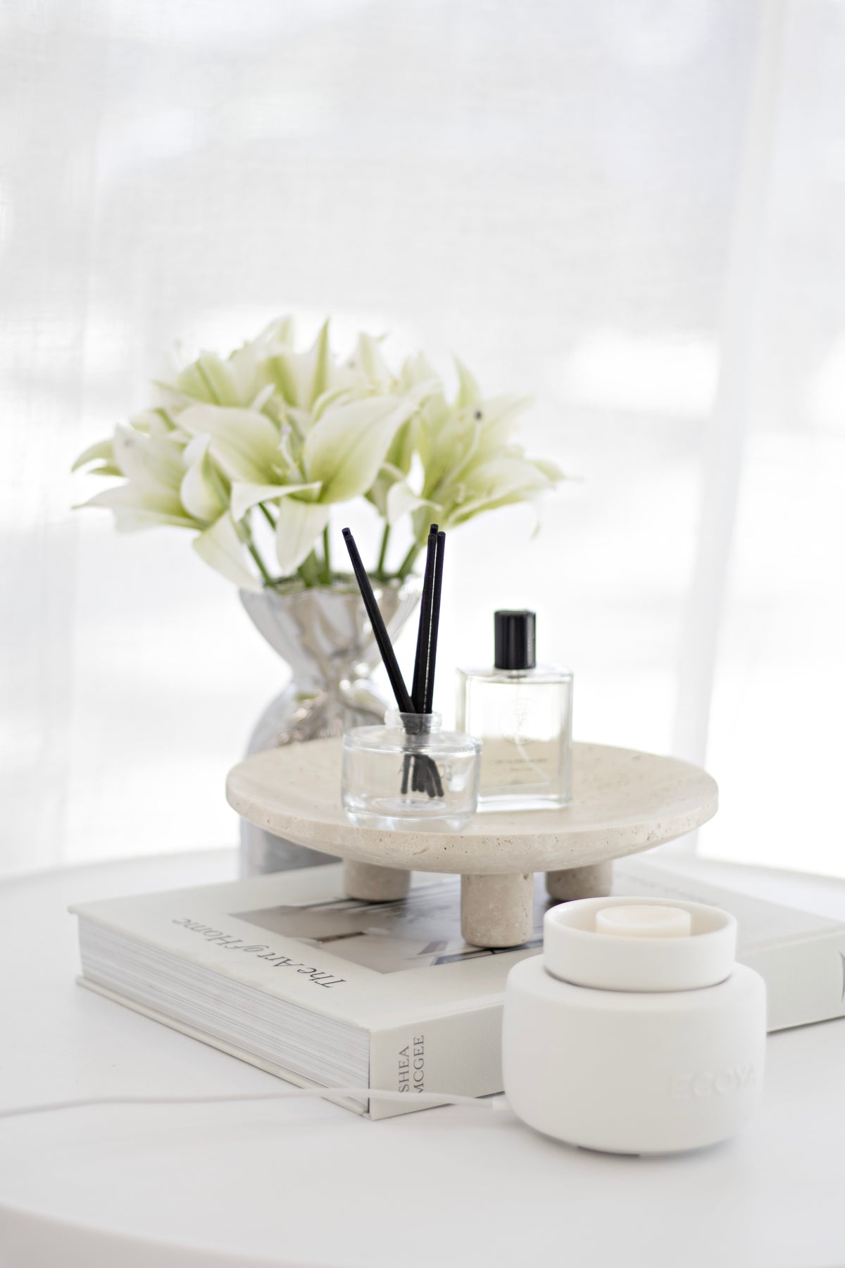 Cream lilies in a vase on a tray with a reed diffuser, candle and stacked books