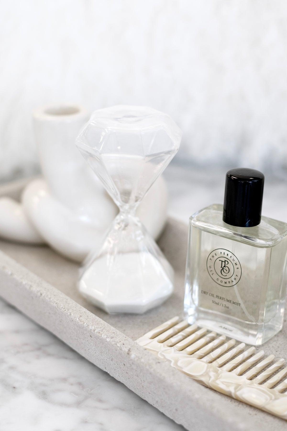 Concrete tray on marble holding a perfume bottle, faceted glass hourglass and comb, knot holder behind